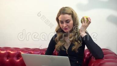 漂亮的金发美女在红色沙发上使用<strong>笔记本</strong>电脑，并在客厅吃一个绿色<strong>苹果</strong>。